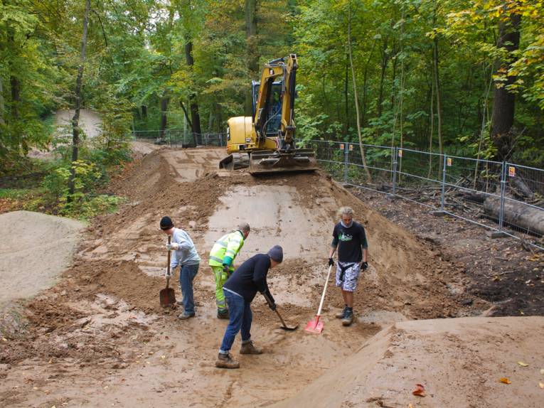 Vier junge Männer bringen eine Mountainbike-Strecke in Form, im Hintergrund wird mit einem Bagger ein Hüggel aufgeschichtet